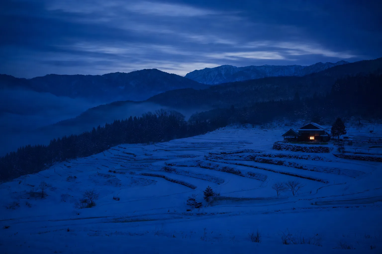 夕暮れの雪に覆われた棚田。遠くの農家に一つだけ灯りがともる